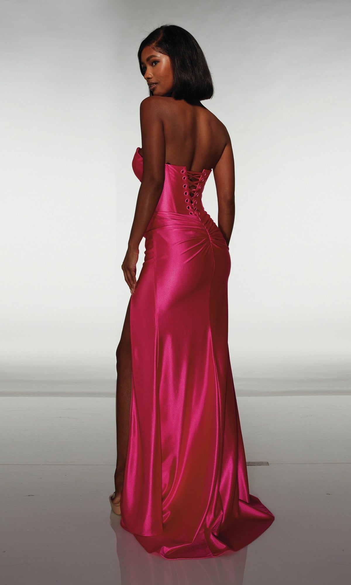A woman faces away from the camera in the Alyce 62046 long prom dress—a strapless, bright pink satin gown with a corset bodice, high-leg slit, and lace-up back.